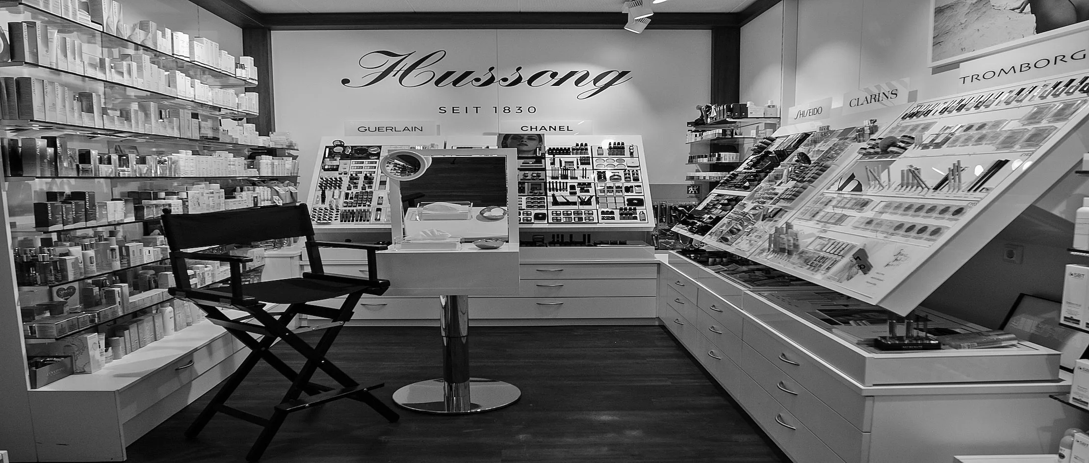 Unser Kosmetikangebot in der Filiale am Mainzer Dom - schwarz-weiß Bild eines Kosmetikladens mit verschiedenen Makeup-Produkten auf Regalen und Ausstellern. Ein Regiestuhl steht in der Mitte. An der Wand steht der Schriftzug Hussong seit 1830. Marken wie Guerlain, Chanel und Clarins sind sichtbar.